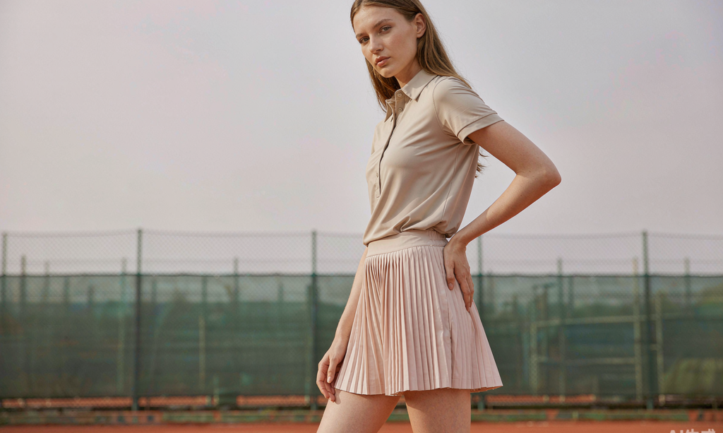 Female model wearing a pleated off-white tennis skort and a beige performance polo on an outdoor clay court, premium editorial activewear photography
